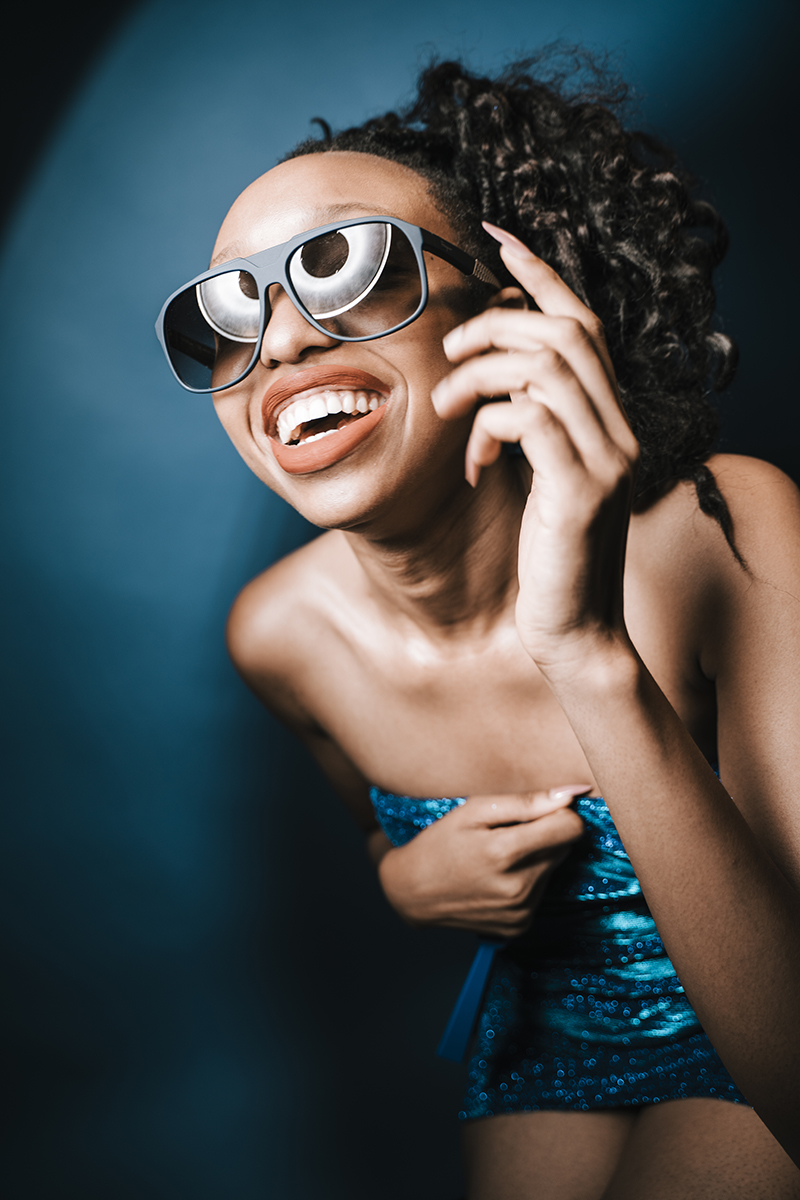 Female model laughing in studio holding onto dress falling off with sunglasses beauty dish reflection