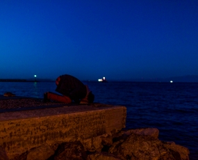 Port of Chios. Falah, a 26 year Pakistani refugee, pray on the harbour. In the background the Turkish coast.